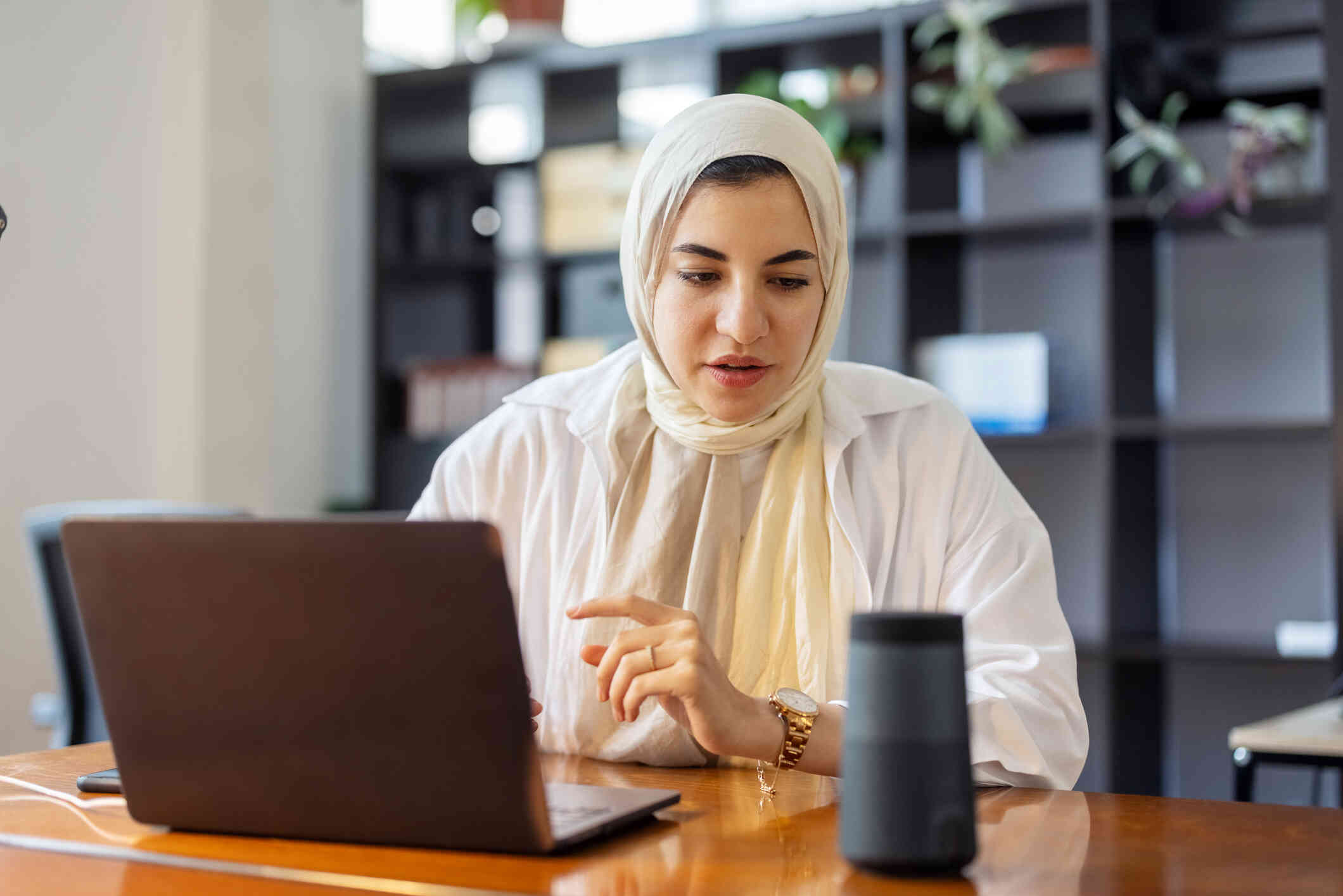 A woman in a tan hijab sits at a table with her laptop open infront of her while talking to the smart device on the table next to her.